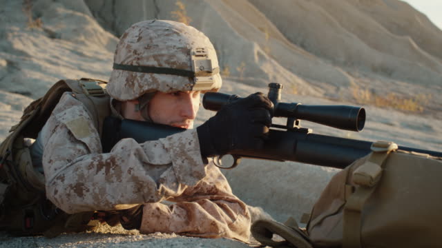 Close-up-of-Sniper-Lies-Down-on-the-Hill-and-Aims-through-the-Rifle-Scope-in-Desert-Environment
