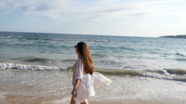 Mujer-feliz-en-bikini-y-camiseta-caminando-por-la-playa-cerca-del-océano-y-levantando-la-mano.-Joven-hermosa-chica-disfrutando-de-la-vida-y-la-diversión-a-la-orilla-del-mar.-Vacaciones-de-verano-o-vacaciones-de-paisaje-en-el-fondo