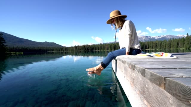 Woman's-feet-dangle-from-wooden-pier,-above-lake