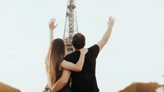 Joven-pareja-feliz-caminar-cerca-de-la-Torre-Eiffel-en-París,-Francia.-Mirada-de-hombre-y-mujer-y-monumento-y-levanta-las-manos