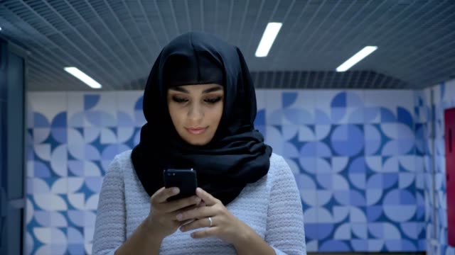 Young-charming-muslim-woman-in-hijab-is-typing-message-on-smartphone-in-underpass,-watching-at-camera,-religion-concept,-communication-concept