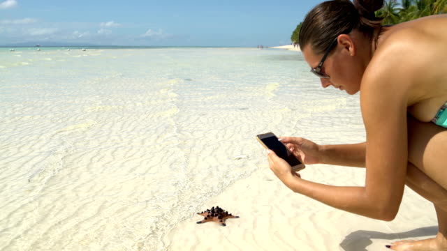 A-woman-in-a-swimsuit-takes-pictures-of-a-starfish-on-a-smartphone