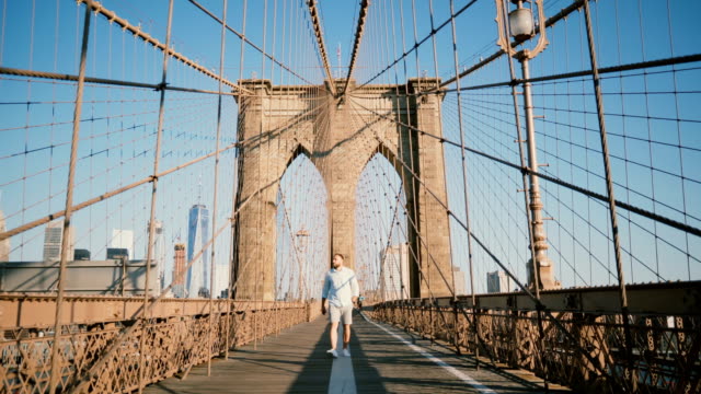 Positive-male-European-tourist-walking-towards-camera,-smiling-and-looking-around-along-Brooklyn-Bridge,-New-York-4K