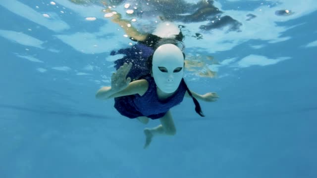 An-unusual-little-girl-swims-and-poses-underwater-in-a-fabulous-white-mask-and-purple-dress,-looks-at-the-camera-and-waving-her-hands-on-a-blue-background.-Slow-motion.