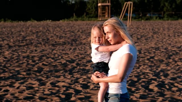 Schöne-blonde-Mutter-und-Tochter-kuscheln-und-sprechen-am-Strand-bei-Sonnenuntergang.