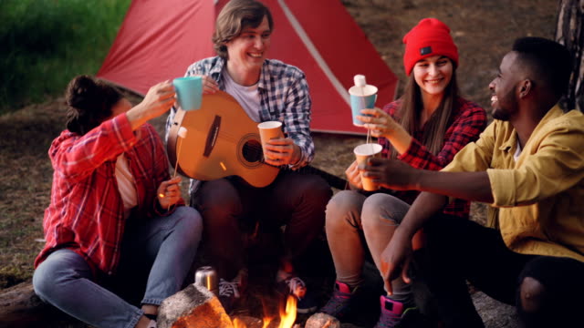 Fröhliche-Junge-Menschen-Freunde-sind-klirrende-Gläser-mit-Getränken-sitzen-um-Feuer-im-Wald-mit-warmen-Marshmallow-auf-Sticks,-Lächelnder-Mann-hält-Gitarre.