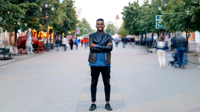 Time-lapse-portrait-of-cheerful-African-American-man-standing-in-city-center-wearing-stylish-clothes-looking-at-camera-and-smiling-while-people-are-passing-by.
