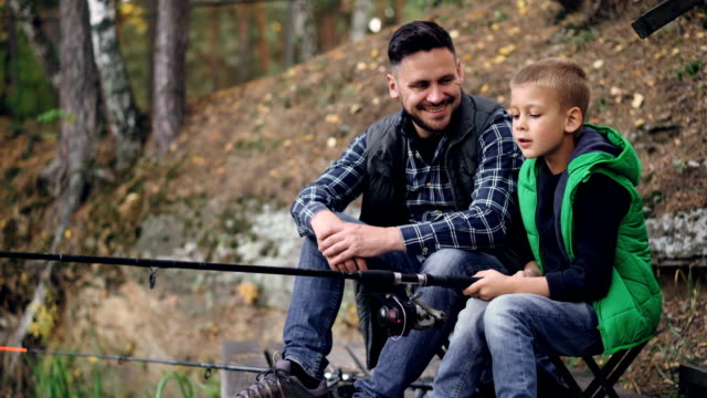 Handsome-guy-loving-dad-is-teaching-his-son-to-catch-fish-sitting-on-river-bank-together,-boy-is-holding-rod-and-moving-it-talking-to-daddy.-Hobby,-adventure-and-family-concept.