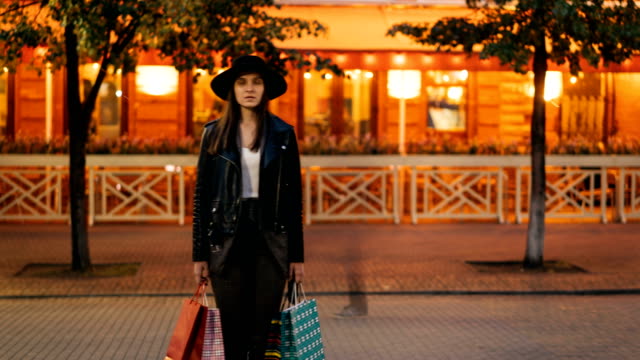 Zoom-out-time-lapse-of-serious-young-lady-in-fashionable-garments-standing-outdoors-in-pedestrian-street-holding-shopping-bags-and-looking-at-camera,-busy-people-are-passing-by.