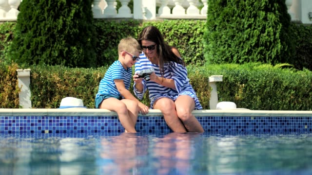 Young-mother-taking-photo-of-little-son-using-professional-camera-on-edge-swimming-pool