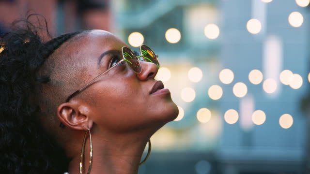 Young-black-woman-wearing-round-sunglasses-looking-up,-side-view,-head-shot,-bokeh-lights-in-background