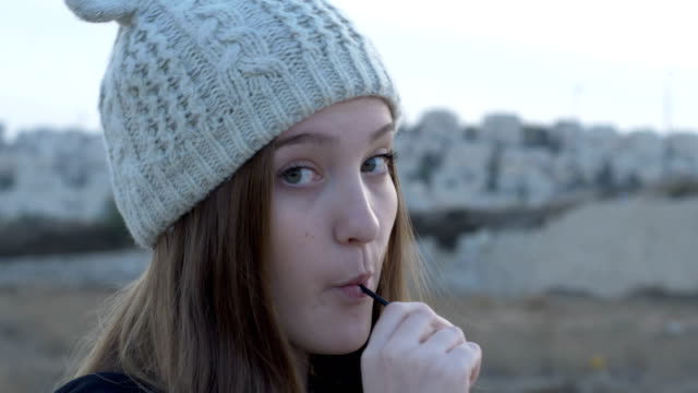 Beautiful-teenage-girl-in-a-gray-knitted-cap,-which-showing-and-eating-lollipop,-artistic-and-expressive-playing-on-camera,-outdoor-footage.