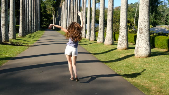 Young-beautiful-woman-jogging-and-spinning-along-exotic-parkland-and-having-fun.-Happy-girl-is-running-in-tropical-park.-Freedom-travel-concept.-Summer-vacation-trip-Rear-back-view