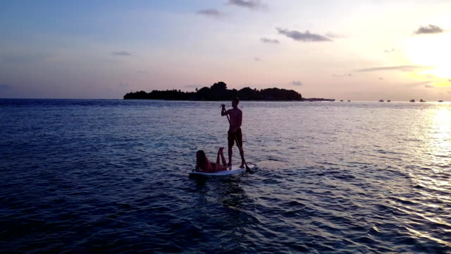 v04104-fliegenden-Drohne-Luftaufnahme-der-Malediven-weißen-Sandstrand-2-Personen-junges-Paar-Mann-Frau-Paddleboard-Rudern-Sonnenuntergang-Sonnenaufgang-auf-sonnigen-tropischen-Inselparadies-mit-Aqua-blau-Himmel-Meer-Wasser-Ozean-4k