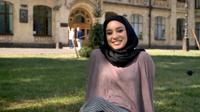Young-happy-muslim-girl-in-hijab-is-sitting-on-lawn-in-sun,-watching-at-camera,-builging-on-background,-religious-concept,-relax-concept