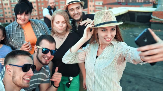 Elegant-young-woman-in-hat-taking-selfie-surrounded-by-friends-using-smartphone-at-rooftop-party