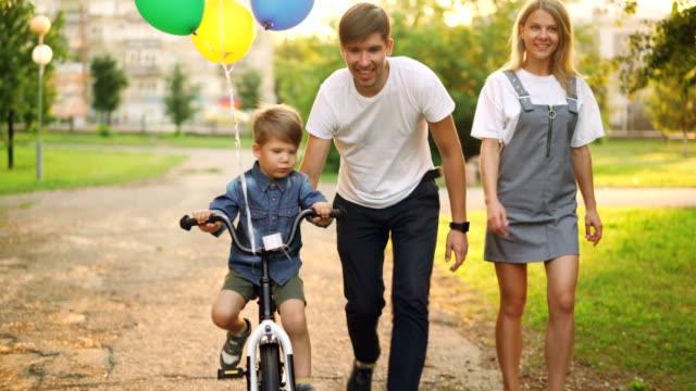 Loving-parents-are-teaching-son-cute-boy-to-ride-bike-in-city-park,-child-is-cycling-and-his-father-is-holding-him-helping-to-balance-while-mother-is-walking-beside-them.