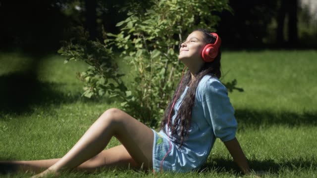 Young-girl-with-long-hair-listening-to-music-on-headphones-using-smartphone-sitting-on-grass-in-park-in-sunny-weather.-4K