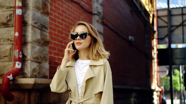 Attraktive-Frau-mit-blonden-Haaren-spricht-auf-dem-Handy-zu-Fuß-entlang-der-Straße-in-modernen-Stadt.-Junge-Dame-trägt-trendige-schwarze-Sonnenbrille-und-Sommerfell.