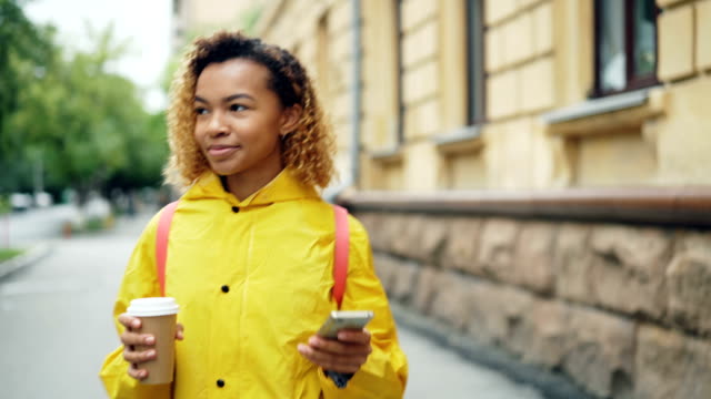 Lächelndes-Afroamerikanisches-Mädchen-nutzt-Smartphone-SMS-Freunde-und-halten-zum-Mitnehmen-Kaffee-zu-Fuß-in-die-Stadt-allein.-Moderne-Technik,-Kommunikation-und-Getränke-Konzept.