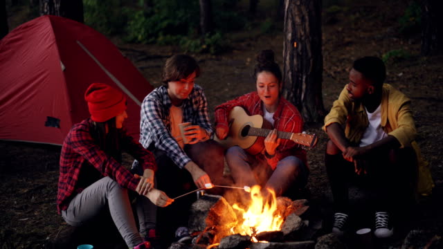 Pretty-woman-is-playing-the-guitar-while-her-friends-are-singing-funny-songs-and-laughing-cooking-marshmallow-on-fire-and-clapping-hands.-Music,-nature-and-fun-concept.