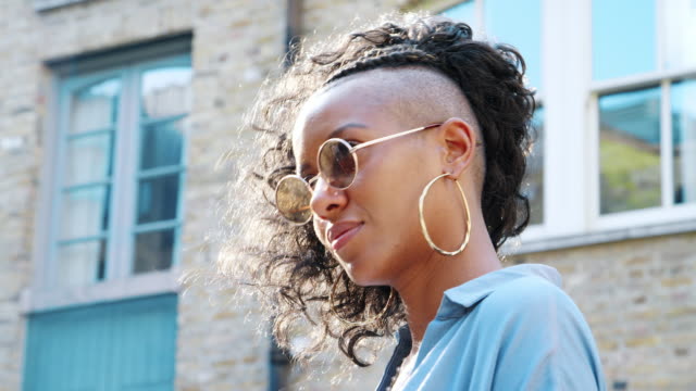 Fashionable-young-black-woman-wearing-blue-dress-and-sunglasses-laughing-to-camera-in-the-street,-backlit,-low-angle,-close-up