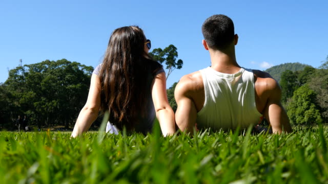 Junges-Paar-am-grünen-Rasen-im-Park-liegen-und-entspannen.-Mann-und-Frau-sitzen-auf-der-Wiese-am-Natur-und-küssen.-Mädchen-und-Jungen-mit-Blick-auf-die-Landschaft-und-Urlaub-genießen.-Hinten-wieder-Low-Blickwinkel