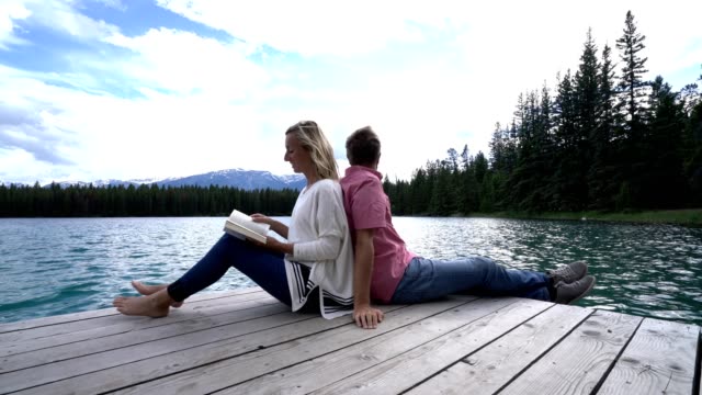 Couple-relaxing-on-wooden-lake-pier,-sitting-back-to-back,-Canada