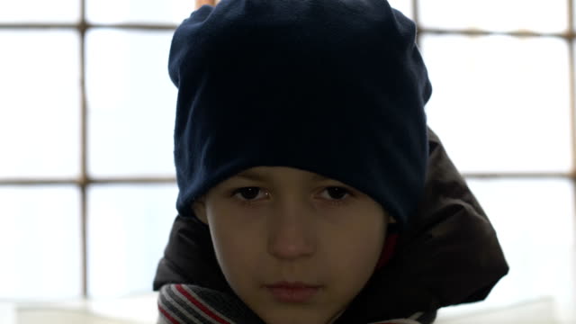 portrait-of-unhappy-boy-stands-alone-near-a-broken-window,-looking-into-the-camera,-cold-weather