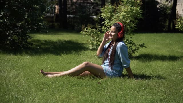 Young-girl-listening-to-music-on-headphones-using-smartphone-sitting-on-grass-in-park-in-sunny-weather.-4K
