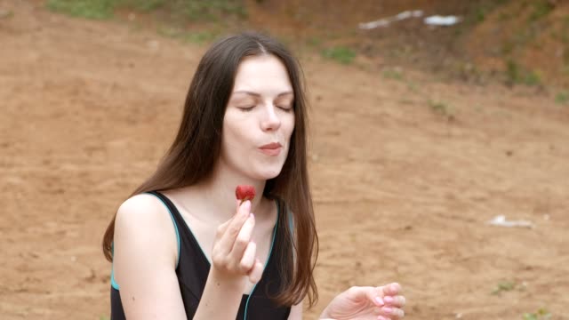 Brünette-junge-Frau-isst-eine-Erdbeere-am-Strand-sitzen.-Seitenansicht.