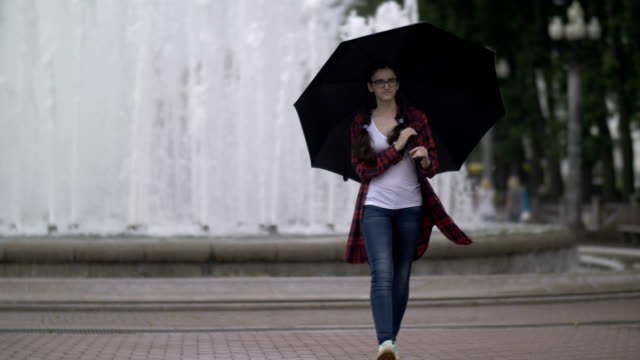 girl-with-an-umbrella-walking-in-the-park-on-a-background-of-a-fountain,-looking-at-the-camera