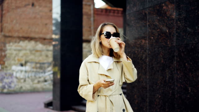 Elegante-junge-Dame-ist-Fuß-entlang-der-Straße,-mit-Smartphone-und-trinken-to-Go-Kaffee-genießen-zu-Fuß-im-schönen-modernen-Stadt.-Menschen,-modernen-Lifestyle-und-Getränke-zum-Mitnehmen-Konzept.