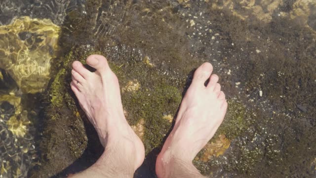 Male-legs-standing-on-stones-in-river-water-point-of-view.-Barefoot-man-in-river