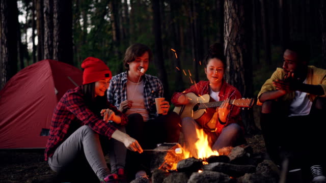 Happy-adventurous-hikers-are-sitting-around-fire,-singing-and-playing-the-guitar,-eating-marshmallow-and-clapping-hands-on-dark-summer-evening.-Tent-in-forest-is-visible.