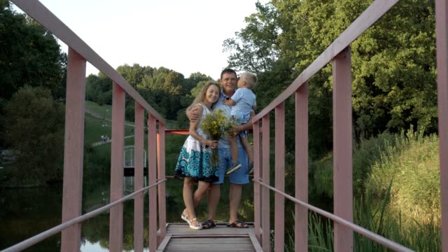 Happy-dad,-mom-and-little-son-are-standing-on-the-bridge-over-the-pond-in-the-Park-against-the-landscape.-The-son-sits-on-the-hands-of-the-Pope,-kisses-and-hugs-him-with-his-mother.