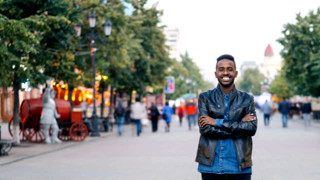 Time-lapse-of-smiling-African-American-man-standing-alone-in-city-center-with-hands-crossed-enjoying-city-life-and-looking-at-camera.-Youth-and-lifestyle-concept.