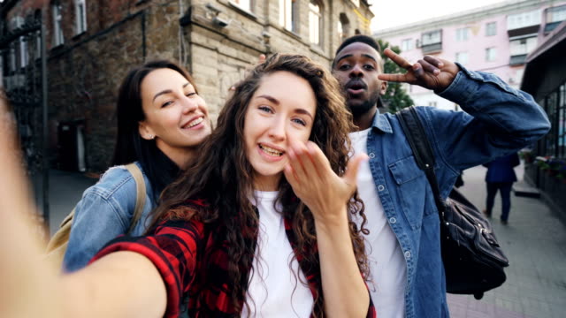 Point-of-view-shot-of-joyful-girls-and-guys-multiethnic-group-taking-selfie-holding-camera-and-posing-outdoors-during-vacation-in-beautiful-city.-Tourism-and-photography-concept.