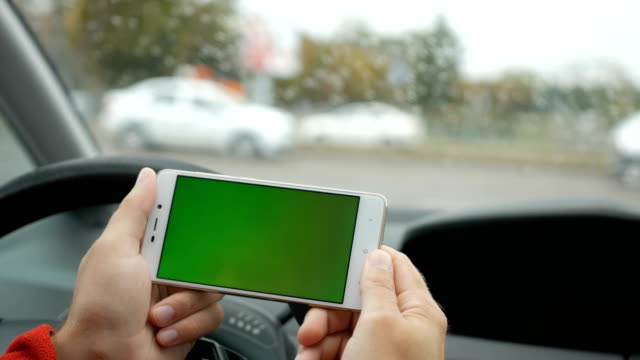 Hand-holding-smartphone-with-green-blank-screen-in-car-for-direction,-massage,-location,-business.-City-life.-Man-sits-in-car-and-works-on-smartphone-green-screen-closeup.-Chroma-key.-Rainy-day.