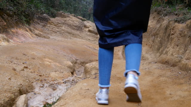 Following-to-female-tourist-in-raincoat-walking-on-stone-mountain-trail.-Feet-of-woman-hiking-through-a-narrow-canyon.-Legs-of-girl-in-sneakers-going-on-the-rocky-road.-Low-angle-of-view-Close-up