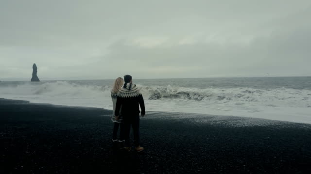 Aerial-view-of-young-couple-standing-on-the-black-volcanic-beach-near-the-troll-toes-rock-and-looking-on-wave-in-Iceland