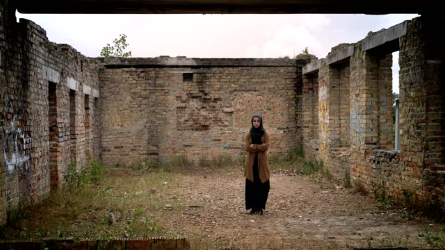 Young-muslim-woman-in-hijab-standing-alone-in-ruined-abandoned-brick-building