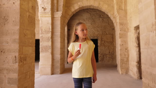 Young-girl-walking-and-eating-ice-cream-on-background-with-arch-from-beige-stones