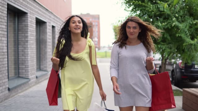 Two-beautiful-young-girls-walk-down-the-street-with-bags-in-their-hands-after-shopping,-having-a-good-mood.-4K
