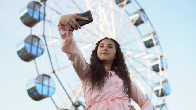 A-girl-with-long-hair-in-a-pink-dress-makes-selfie-using-a-smartphone-standing-near-the-Ferris-wheel.-4K