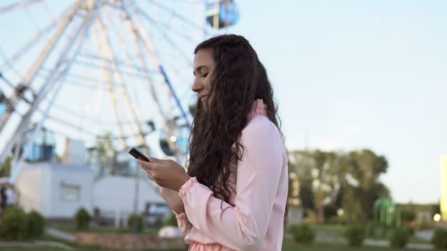 The-girl-walks-by-the-ferris-wheel-and-uses-a-smartphone.-4K