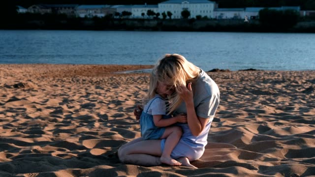 Schöne-glückliche-blonde-Mutter-und-Tochter-sprechen-bei-Sonnenuntergang-am-Strand-sitzen-und-Kuscheln.