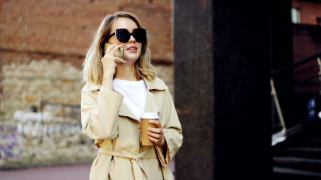 Beautiful-young-woman-with-blond-hair-is-talking-on-mobile-phone-and-drinking-take-away-coffee-walking-in-city-along-street-and-enjoying-drink-and-conversation.