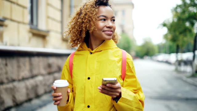 Happy-African-American-woman-is-using-smartphone-touching-screen-and-smiling-walking-outdoors-in-beautiful-city-with-to-go-coffee.-Modern-lifestyle-and-communication-concept.