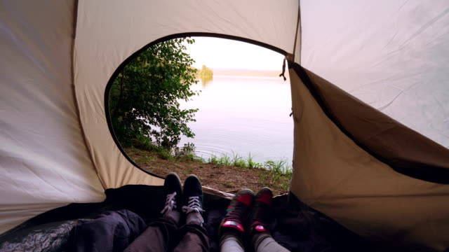Touristen-liegen-im-Zelt-machen-Tanzbewegungen-Spaß-entspannte-Füße-zu-bewegen,-ist-die-schöne-Aussicht-auf-See-oder-Fluss-außerhalb.-Mensch,-Natur-und-camping-Konzept.